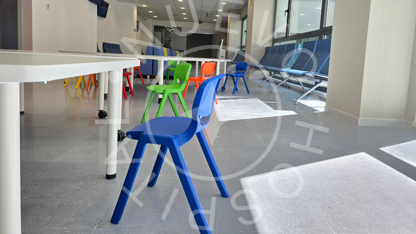 En primer plano sillas pequeñas de colores y mesa blanca y al fondo a la derecha sillas de sala de espera azules