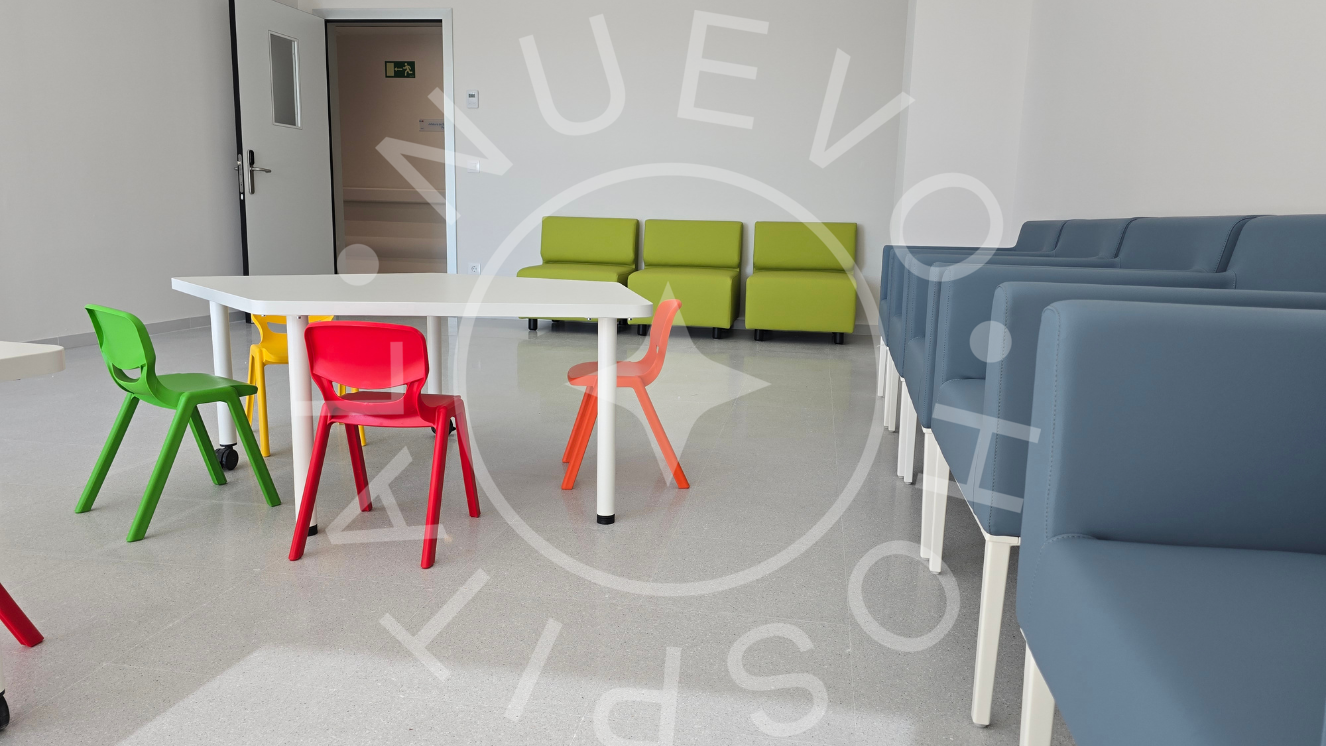 Sala de juegos hospitalización pediátrica con sillones de espera azules a la derecha y a la izquierda mesa con sillas de colores pequeñas y al fondo sillones verdes