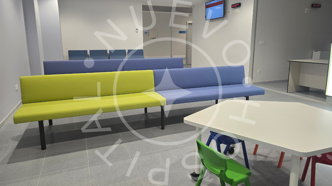 Sillones corridos en sala de espera de color verdoso y azul y en primer plano mesa blanca con sillas de colores pequeñas