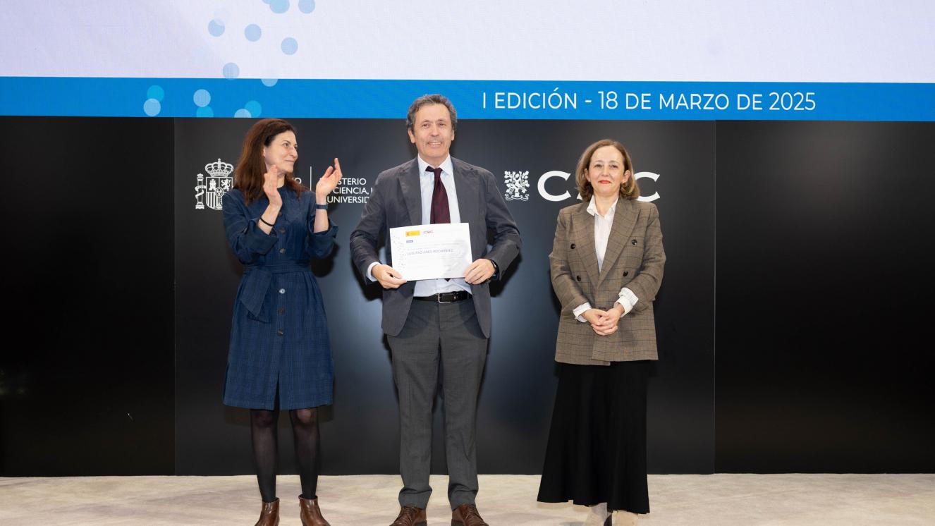 Luis Paz-Ares recogiendo Diploma Ramón y Cajal CSIC en escenario entre dos mujeres