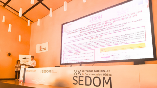 Juan Martín Romero en atril al fondo y a la izquierda de gran pantalla con texto