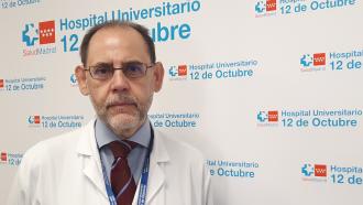 Retrato de hombre con barba blanca recortada y gafas con camisa, corbata y bata blanca sobre fondo de logos