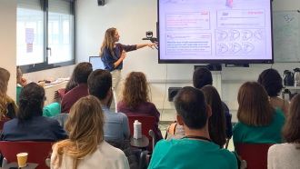 Aula vista desde atrás con alumnos adultos y profesora delante señalando una pantalla digital
