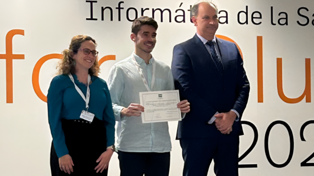 Tres personas posando en escenario, el del medio sosteniendo un diploma
