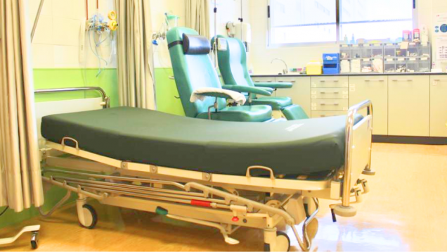 Una cama de hospital en primer plano y dos sillones detrás, verdes, y al fondo armarios y una ventana