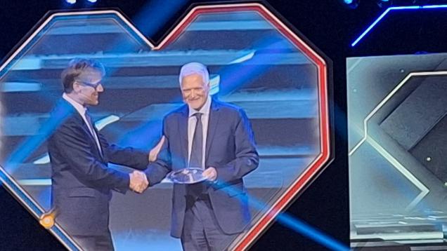 Auditorio a oscuras y ocupando gran parte de la imagen la pantalla donde se ve a un hombre con traje azul recibiendo un premio y apretando la mano de otro hombre con traje azul