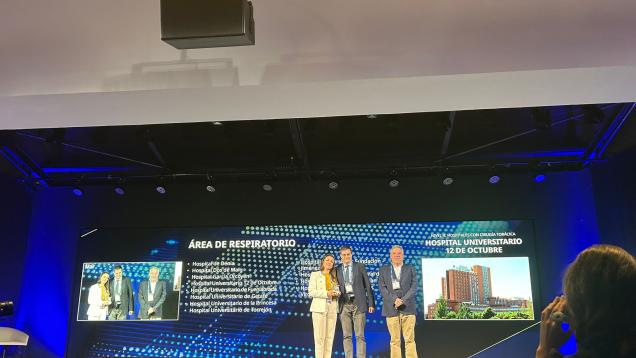 Tres personas en un escenario dos hombres a la derecha y una mujer a la izquierda  con un premio 
