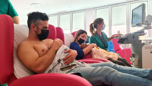 Cuidado piel con piel un padre en primer plano con bebé sobre su pecho al descubierto y en segundo plano una mamá con su bebé también sobre su pecho