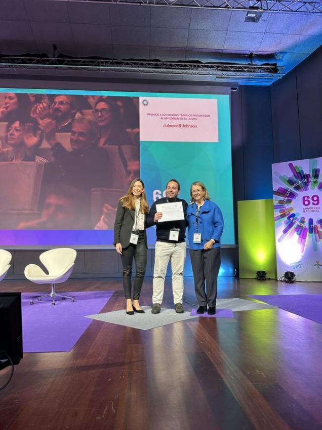 Foto de tres personas en escenario en el medio hombre con diploma en las manos