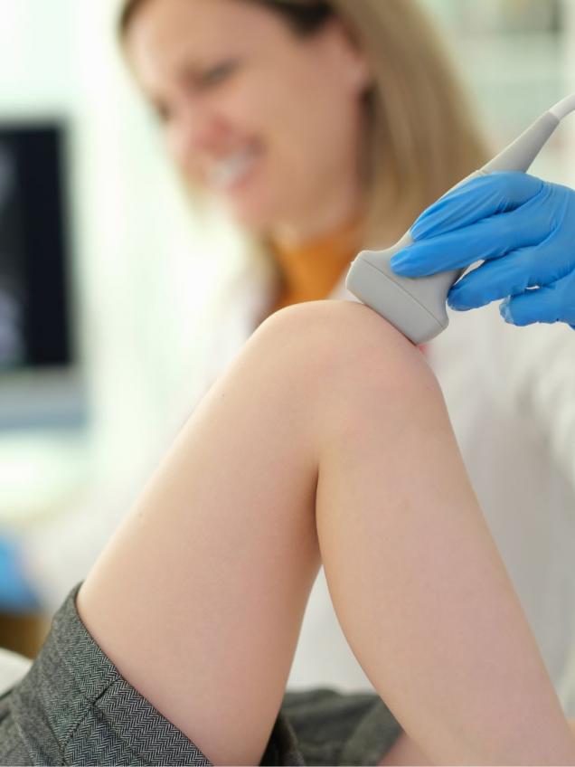 una doctora a la derecha hace una ecografía en la rodilla a una niña tumbada ala izquierda